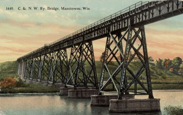 View from shoreline toward a railroad bridge on the right crossing high above the Manitowoc River. Caption reads: "C. & N.W. Ry. Bridge, Manitowoc, Wis."
