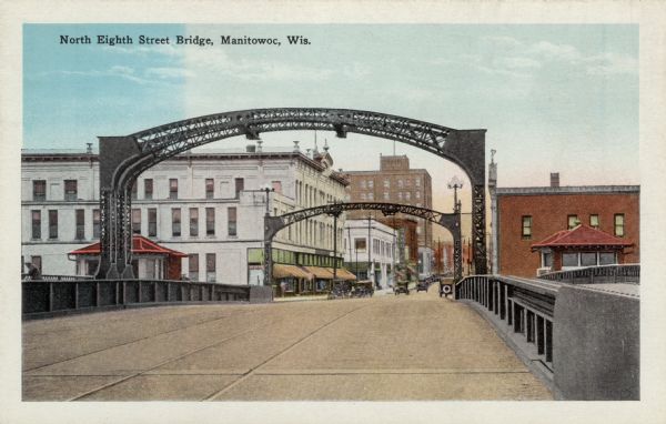 View from street bridge leading into downtown Manitowoc. Automobiles are parked at the curbs, and streetcar tracks are running down the center of the street. Caption reads: "North Eighth Street Bridge, Manitowoc, Wis."