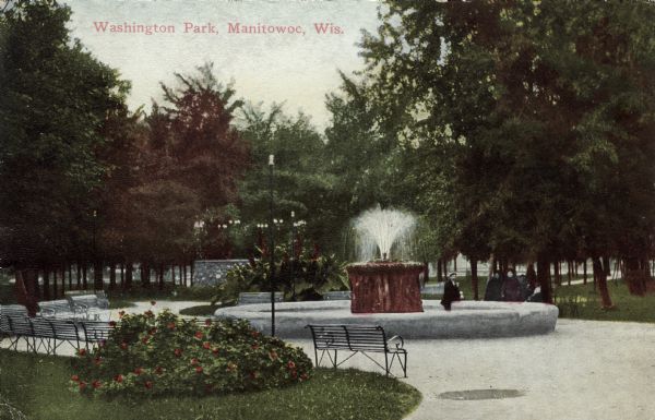 View of the fountain in Washington Park. A group of people are next to the fountain. Park benches are bordering the walkway. Caption reads: "Washington Park, Manitowoc, Wis."