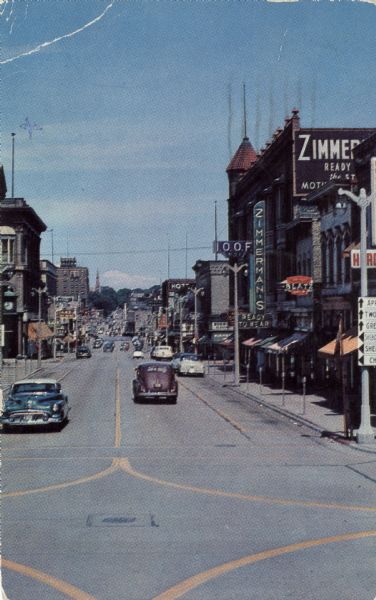 Slightly elevated view of 8th Street, looking north in a central business district. Cars are parked along the curb, and other cars are moving down the street. There is clothing store called Zimmerman's on the right.