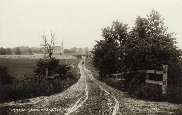 View down a long dirt road. In the distance the road passes under a railroad bridge near a church with a cemetery. Caption reads: "Lover's Lane, Mapleton, Wis."