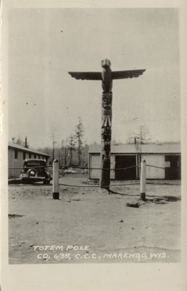 View of a totem pole at a Civilian Conservation Corps camp. An automobile parked next to barracks in the background.