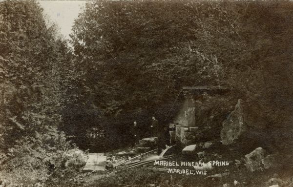 View toward two men standing near a mineral spring in the woods. Caption reads: "Maribel Mineral Spring, Maribel, Wis."