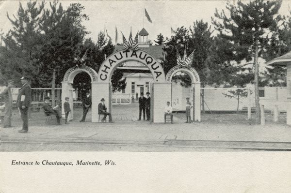 View towards people sitting and standing near the front gates to the Chautauqua. Flags are on the arches. Caption reads: "Entrance to Chautauqua, Marinette, Wis."