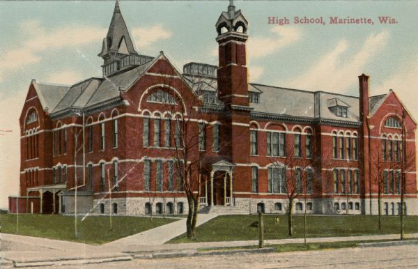 View across road toward the high school building. A dog is standing on the lawn. Caption reads: "High School, Marinette, Wis."