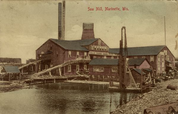 View of a sawmill next to a river. Conveyor ramps are leading into the building. Caption reads: "Saw Mill, Marinette, Wis."