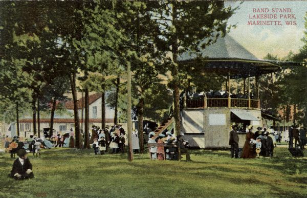 View of a bandstand at a park. A band is playing, and people are gathered on the grounds of the park. Caption reads: "Band Stand, Lakeside Park, Marinette, Wis."