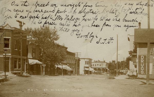 View of Main Street, with a bank building on the left and a drugstore on the right. Horse-drawn vehicles are along the curb on the right in the background. Caption reads: "Main St. Marion, Wis."