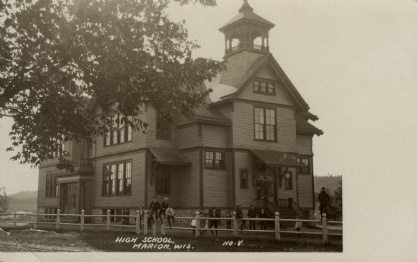 Exterior view of a two-story wood-frame school. Students are standing near the door and sitting on the fence. Caption reads: "High School, Marion, Wis."