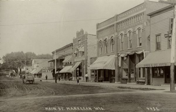 View across Main Street toward businesses on the right, including a harness repair shop, undertaker, and a bank. A horse and wagon is in the street. Caption reads: "Main St. Markesan, Wis."