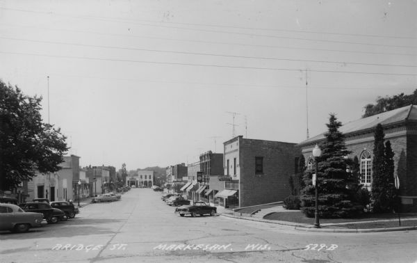 View down Bridge Street which is lined with businesses. Automobiles are parked at an angles along the curbs. Businesses include an electronics (TV/radio) store, a bank, a bar, a theater, and a service station at the far end of the street. Caption reads: "Bridge St., Markesan, Wis."