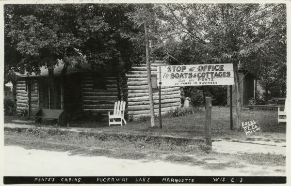 Exterior view of a vacation log cabin for Pentz Boats and Cottages. There is an outhouse in the back under a tree. Caption reads: "Pentz's Cabins, Puckaway Lake, Marquette, Wis."