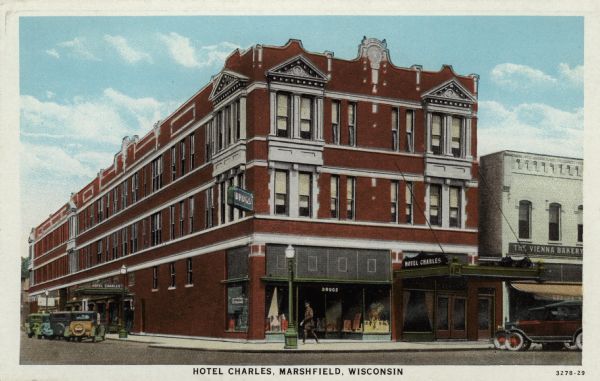 Exterior view of the Hotel Charles, at the corner of Second and Main Street. A drug store is on the corner, with a bakery next door. Automobiles are parked at the curbs. Caption reads: "Hotel Charles, Marshfield, Wis."