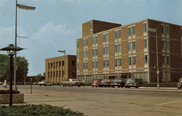 Color postcard of the exterior of Marshfield Clinic. Automobiles are parked at the curb at parking meters.
