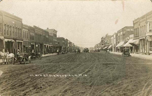View looking down unpaved Main Street which is lined with businesses. A billiard hall and millinery are on the left; a hotel is on the right. Horse-drawn vehicles and automobiles are in the street, and pedestrians are on the sidewalk. Caption reads: "Main St., Marshfield, Wis."