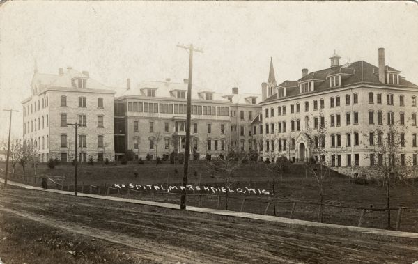 View of a four-story hospital on a dirt road. There is a wooden plank sidewalk along road. Caption reads: "Hospital, Marshfield, Wis."