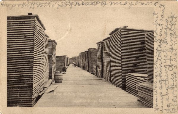 View of rows of stacked wooden palettes. A wood plank sidewalk is between them. Caption reads: "Row of Wis. Timber & Land Cos., Lumber Piles, Mattoon, Wis."