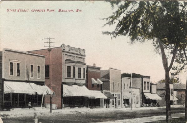 View of a block of storefronts, many with awnings over the windows. Caption reads: "State Street, Opposite Park, Mauston, Wis."