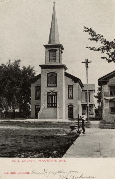 View of the M.E. Church. A boy is standing next to a water pump on the side of the street. Caption reads: "M.E. Church, Mauston, Wis."