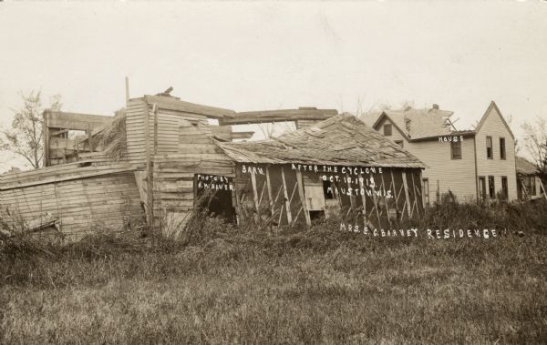 View of a collapsed barn after a tornado. The house in the background on the right is missing its roof. Captions read: "Barn," "House," "After the Cyclone, Oct. 10, 1913, Mauston, Wis." and "Mrs. E.G. Barney Residence."