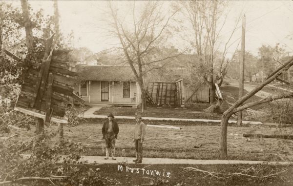 View looking down towards two men standing on a sidewalk in a residential neighborhood after a tornado. There are damaged trees, and part of a wooden wall is stuck in a tree on the left. There is a house across the street and another large piece of a wooden wall is lying in the yard. Caption reads: "Mauston, Wis."