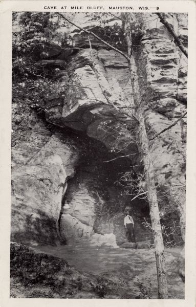 View of a cave in a bluff, with a man standing in the entrance. Caption reads: "Cave at Mill Bluff, Mauston, Wis."