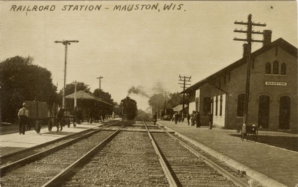 View down a set of two railroad tracks towards the Mauston Railroad Station on the right, and an approaching train on the left set of tracks. Workers and passengers are on the platform on the left, and also on the right near the station. Caption reads: "Railroad Station -- Mauston, Wis.