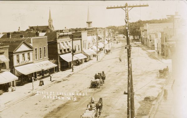 Elevated view of town. Horse-drawn vehicles are on State Street. There is a sign for "Billiards" on a building in the right foreground. Other storefronts line the left side of the street with signs in front, including "Furniture/Undertaking," "S.B. Mann Restaurant Hotel," and "E.F. Smith." A large church is in the background.