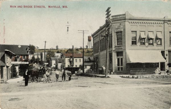 View of the intersection of Main and Bridge Streets. A dentist's office on the corner on the right. A group of boys are standing near a fountain in the middle of the street, with one boy standing up on the fountain. Another boy is riding a tricycle near people in a horse-drawn wagon. In the distance is a water tower on a hill. Caption reads: "Main and Bridge Streets, Mayville, Wis."