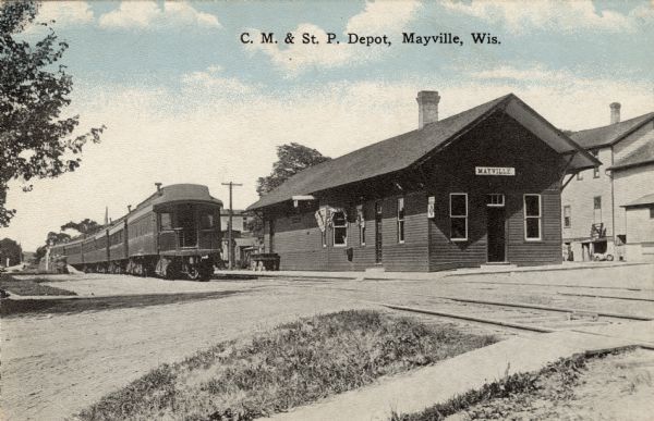 View from road towards the Mayville train depot, with a passenger train on the left set of two railroad tracks. Caption reads: "C.M. & St. Paul Depot, Mayville, Wis."