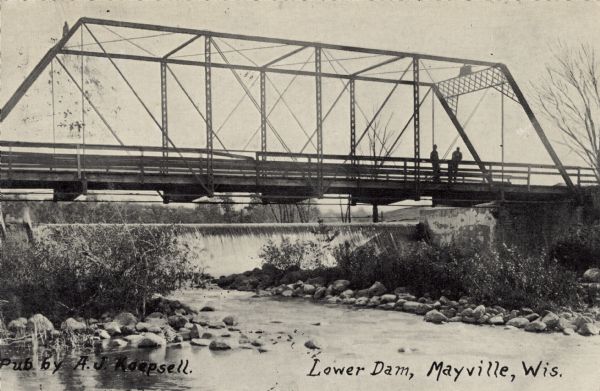 View from shoreline looking toward a dam and bridge on the Rock River near Mayville. Two men are standing on the bridge. Caption reads: "Lower Dam, Mayville, Wis."