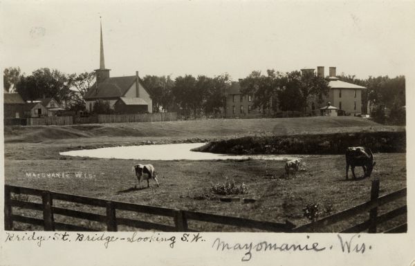 View across fence towards a pasture with cows. A stream is running through the pasture, and in the background behind a fence are buildings and a church among trees. Handwritten caption reads: "Bridge St. Bridge - Looking S.W., Mazomanie, Wis."