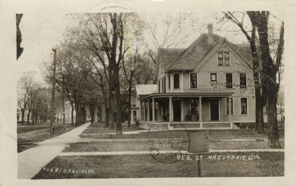 View down the sidewalk and front yards on the right side of a residential street lined with trees and houses. Caption reads: "Res. St. Mazomanie, Wis."
