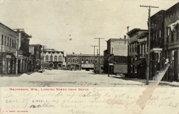 View down the center of a street in the central business district. A dentist's office is on the right. Caption reads: "Mazomanie, Wis., Looking North from Depot."