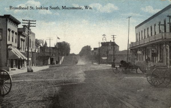 View down center of unpaved street lined with businesses. Horses and wagons are parked along the curbs. Caption reads: "Broadhead St. looking South, Mazomanie, Wis."