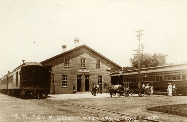 View across street towards the C.M. & St. Paul Depot, with passenger trains on railroad tracks on both sides of the depot. There are pedestrians and a horse-drawn wagon in the front of the depot. Caption reads: "'C. M. & St. P. Depot, Mazomanie, Wis."