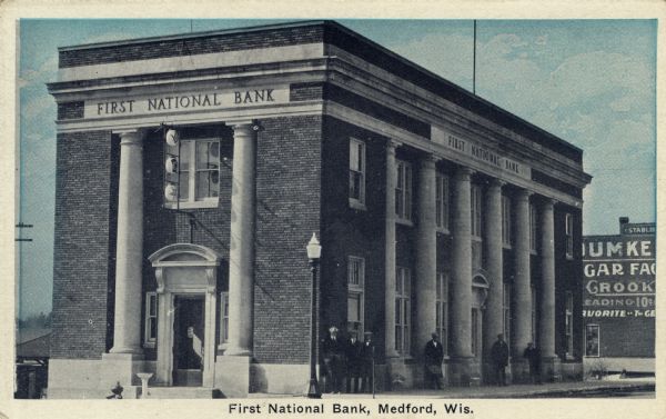 View from street towards the facade of the Neo-Classical bank with columns. A group of men are standing along the right side of the brick building. Caption reads: "First National Bank, Medford, Wis."
