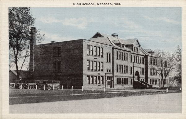 Exterior view of Medford High School. Automobiles are parked on the side of the building. Caption reads: "High School, Medford, Wis."