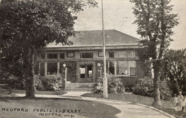 Exterior view of the Medford Public Library. The entrance is flanked by lampposts. A group of girls are standing on the far right. Caption reads: "Medford Public Library, Medford, Wis."
