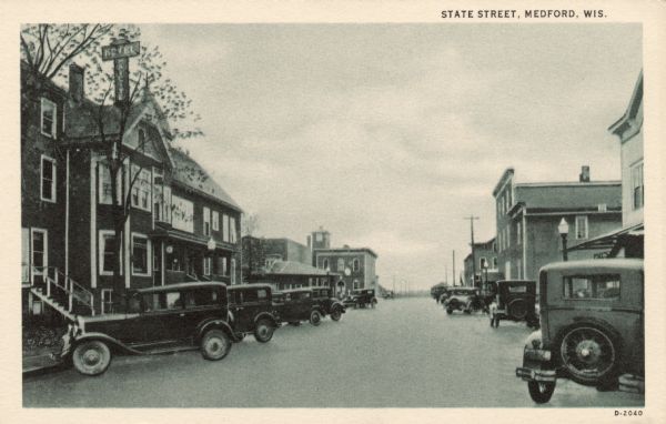 View of a city street lined with automobiles. A hotel is on the left. Caption reads: "State Street, Medford, Wis."