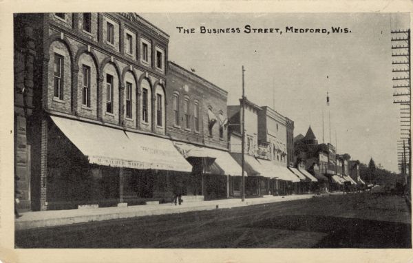 Diagonal view looking left across a street in a business district, including a bakery. Horses and carriages are further down the street. Caption reads: "The Business Street, Medford, Wis."