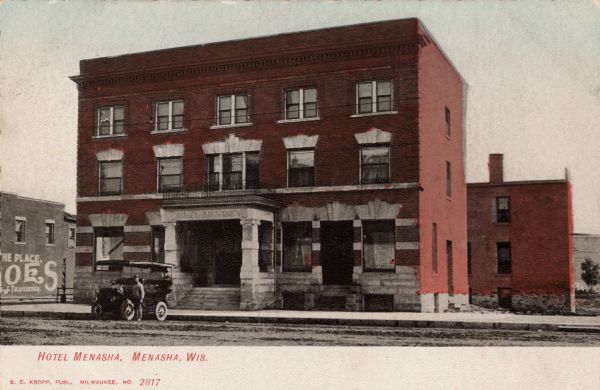 Exterior view of the Hotel Menasha. There is a car and driver at the curb. Caption reads: "Hotel Menasha, Menasha, Wis."