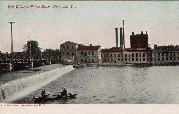 View of a dam on the Fox River and a paper mill on the far bank. Two men are in boat in the foreground. Caption reads: "Dam & Island Paper Mill, Menasha, Wis."