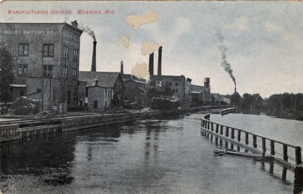 View of factories along the Fox River. The building on the left has a sign painted on it that reads: "Valley Knitting Co." Caption reads: "Manufacturing District, Menasha, Wis."
