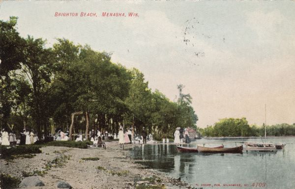View along shoreline of a bathing beach with many people on and near a pier. There are rowboats in the water on the right. Caption reads: "Brighton Beach, Menasha, Wis."