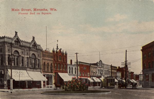 View of a city street lined with businesses. A streetcar is at the intersection, and a  flower bed is in the center of the square. Caption reads: "Main Street, Menasha, Wis. Flower Bed on Square."