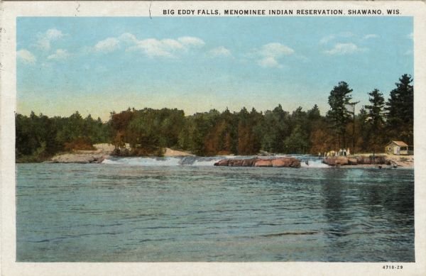 View upriver across water towards the Big Eddy Falls on the Wolf River. A group of people are standing on the right bank. Caption reads: "Big Eddy Falls, Menominee Indian Reservation, Shawano, Wis."