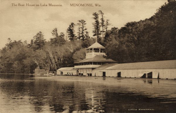 View across lake toward a large multi-level boat house on the shoreline. Caption reads: "The Boat House on Lake Menomin. Menomonie, Wis."