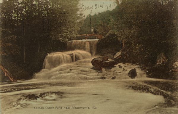 View looking up toward a waterfall with a footbridge spanning the top. A man is on the bridge. Caption reads: "Lambs Creek Falls near Menomonie, Wis."