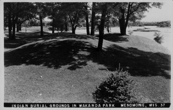 View of burial mounds in a park on the edge of a lake. Caption reads: "Indian Burial Grounds in Wakanda Park, Menomonie, Wis."